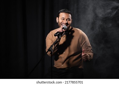 happy indian comedian sitting on chair and performing stand up comedy into microphone on black with smoke