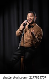 Happy Indian Comedian Sitting On Chair And Performing Stand Up Comedy Into Microphone On Black