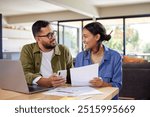 Happy indian couple discussing home finance while holding documents and working on laptop and mobile phone. Middle eastern man and casual woman sitting at table and discussing expenses.