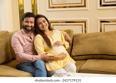 Happy indian couple expecting a baby while husband putting his hand on wife his pregnant wife belly both sitting on couch spending time together. healthcare concept - Powered by Shutterstock