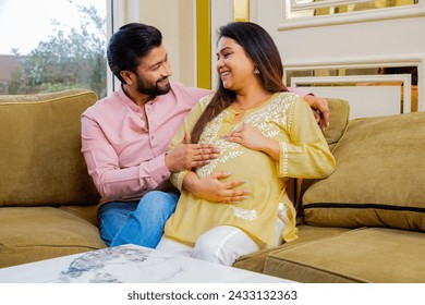 Happy indian couple expecting a baby while husband putting his hand on wife his pregnant wife belly both sitting on couch spending time together. healthcare concept - Powered by Shutterstock