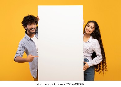 Happy indian couple in love standing by white empty board for advertisement or text over yellow studio background. Excited man and woman standing next to blank placard for ad - Shutterstock ID 2107608089