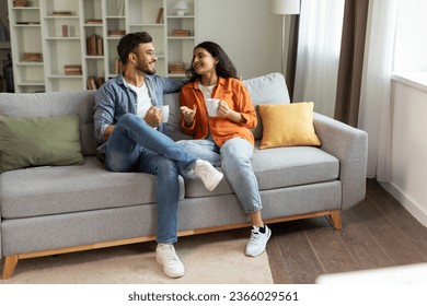 Happy indian couple relaxing and drinking coffee, sitting on couch at home, talking while resting with hot drinks in cups at modern living room, free space - Powered by Shutterstock