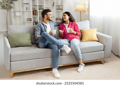 Happy indian couple relaxing and drinking coffee, sitting on couch at home, talking , free space - Powered by Shutterstock