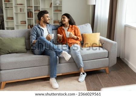 Happy indian couple relaxing and drinking coffee, sitting on couch at home, talking while resting with hot drinks in cups at modern living room, free space