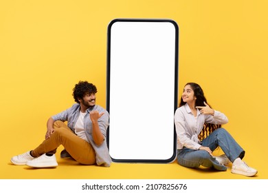 Happy indianisches Ehepaar sitzend nahe dem großen Smartphone mit weißem Bildschirm, demonstriert Kopienraum für App oder Werbedesign, posiert auf gelbem Hintergrund, spitzt Banner auf