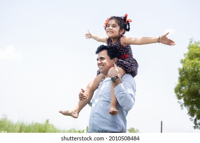 Happy Indian Cute Little Adorable Daughter Sitting On Father Shoulder Having Fun In Field, Spend Leisure Time Together In Farm, Girl Child Enjoy Outdoor, Single Parent, Relationship Bonding 