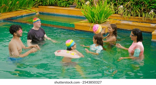 Happy Indian extended family group standing in swimming pool play ball enjoy have fun joy together outdoor home. Smiling older senior parents spend day time with cute teen sibling in hotel resort park