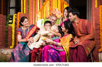 Happy Indian Family Celebrating Ganesh Festival Or Chaturthi - Welcoming Or Performing Pooja And Eating Sweets In Traditional Wear At Home Decorated With Marigold Flowers