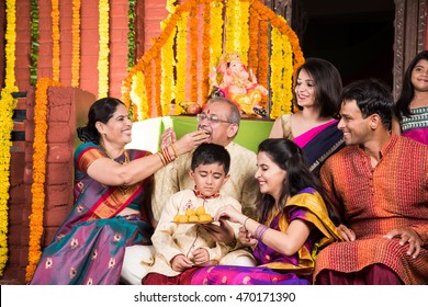 Happy Indian Family Celebrating Ganesh Festival Or Chaturthi - Welcoming Or Performing Pooja And Eating Sweets In Traditional Wear At Home Decorated With Marigold Flowers