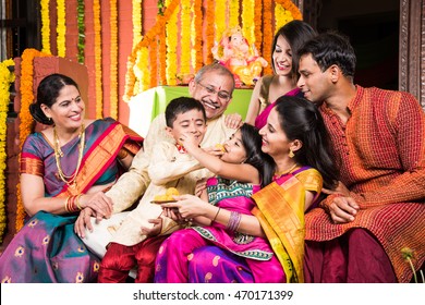 Happy Indian Family Celebrating Ganesh Festival Or Chaturthi - Welcoming Or Performing Pooja And Eating Sweets In Traditional Wear At Home Decorated With Marigold Flowers