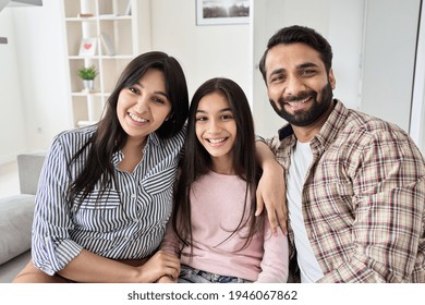 Happy Indian Family Couple And Teen Child Daughter Bonding, Hugging And Smiling Sitting On Couch At Home. Smiling Husband And Wife Embracing Child Looking At Camera Relaxing On Sofa, Portrait.