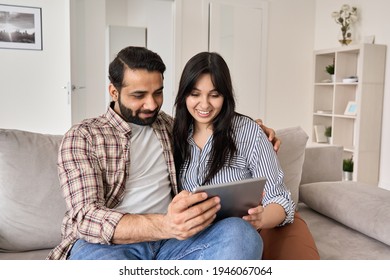 Happy indian family couple using digital tablet computer at home. Smiling young husband and wife watching tv, video calling or doing online ecommerce shopping together sitting on couch in living room.