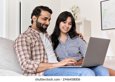 Happy Indian Family Couple Using Laptop Computer Together At Home. Smiling Ethnic Husband And Wife Browsing, Surfing, Doing Online Shopping Or Watching Movie On Pc Device, Hugging Sitting On Couch.