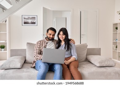 Happy Indian Family Couple Using Laptop Looking At Computer Sitting On Sofa Together Relaxing At Home. Smiling Ethnic Husband And Wife Doing Online Ecommerce Shopping On Website In Modern Living Room.