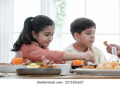 Happy Indian Family Enjoy Eating Food With Hands, Selective Focus On South Asian Cute Girl Holding Naan Bread Dipping In Curry, Wear Traditional Clothes, Sitting With Brother At Dining Table At Home