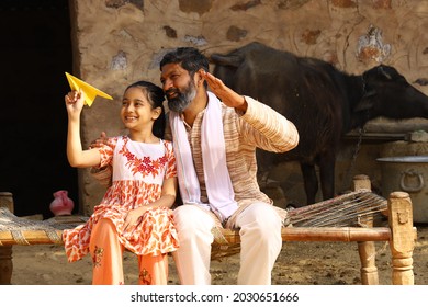 Happy Indian Family Of Father And Daughter Sitting Outdoors On A Folding Bed. Daughter Holding Paper Airplane In Hand.