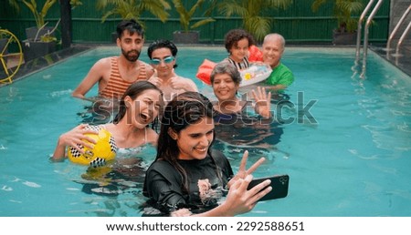 Happy Indian family having fun and enjoying outdoor picnic. Young girl taking selfie with senior parents using mobile smartphone at water park. Cute Child relax in colorful toy floating ring at resort