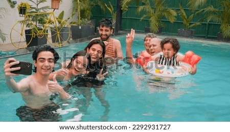 Happy Indian family having fun and enjoying outdoor picnic. Teen boy taking selfie with senior parents using mobile smartphone at water park. Cute Child relax in colorful toy floating ring at resort