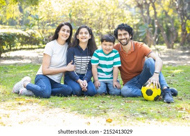 Happy Indian Family Sitting Together In A Summer Park. Young Couple With Their Kids Wearing Casual Cloths Smiling, Enjoying Picnic Holiday Outdoor.