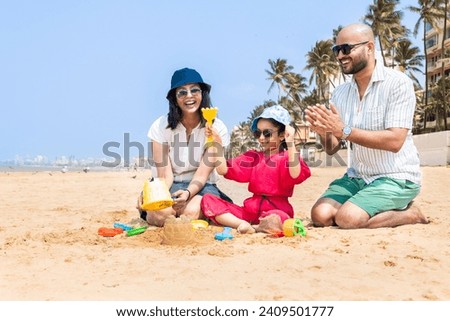 Happy indian family of three making sand castle or home with using sand plastic toys at the tropical beach enjoying holiday, vacation.