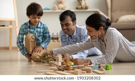 Happy indian family young mom dad and little kid boy spend weekend together play learning game. Caring mixed race parents lying on warm heated floor building dino park from bricks with preschooler son