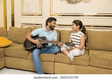 Happy indian father teaching her daughter guitar lessons while sitting on sofa. Child Dream Concept. - Powered by Shutterstock