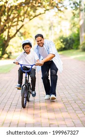 Happy Indian Father Teaching His Son To Ride A Bicycle At The Park