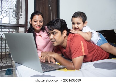 Happy Indian Father With Wife And His Cute Toddler Daughter Lying On Back Using Laptop Surfing Watching On Screen. Family Spend Leisure Time Having Fun Together Looking Something On Computer Display