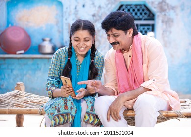 Happy Indian Father And Young Daughter Using Smartphone  While Sitting On Traditional  Bed Outside Their House, Happy Rural Family, Man Point Finger At Mobile Screen. Video Call, Learning Technology 