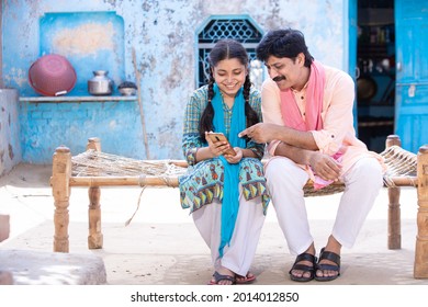 Happy Indian Father And Young Daughter Using Smartphone  While Sitting On Traditional  Bed Outside Their House, Happy Rural Family, Village Man Learning Mobile Phone Technology, Copy Space. 