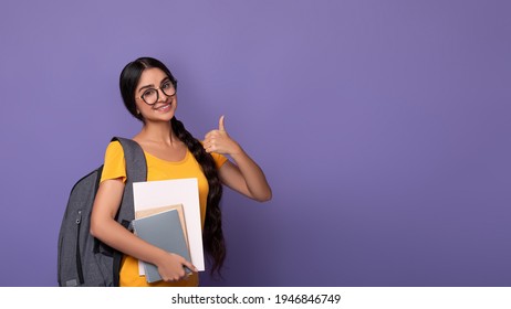 Happy indian female student wearing glasses showing thumbs up sign gesture, holding college materials and wearing backpack, standing isolated on purple studio background with free copy space, banner - Shutterstock ID 1946846749