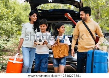 Happy indian fmaily with kids with picnic basket and travel lagguage at destination - concept safe car travel, transportation and vacation or holidays.