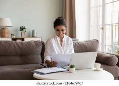 Happy Indian freelance employee woman reading paper document at laptop, smiling at good news, sales report, job result, contract, deal conditions, sitting on couch at laptop, working at home