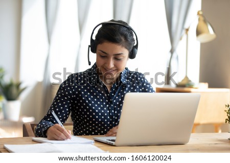 Happy indian girl student wear headset study online with webcam teacher write notes, happy young woman listen lecture watch webinar on laptop sit at desk, distance e learn language education concept