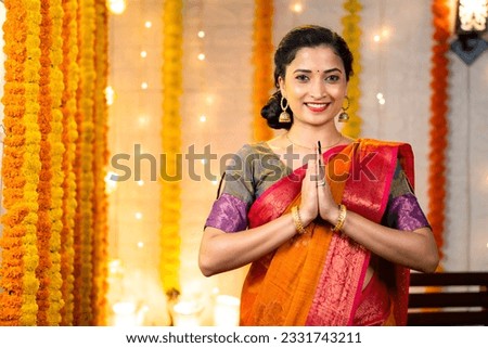 Happy indian girl with traditional ethnic dress showing namaste gesture by looking at camera during festival celebration at home - concept of welcome, greetings and Indian culture.