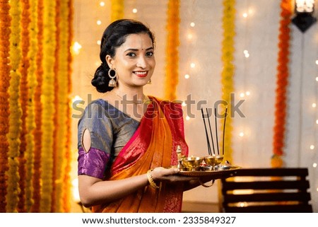 Happy indian girl in traditional ethnic wear holding pooja plate with incense sticks looking camera during diwali festival celebration at home - concept Indian culture, positive emotion and happiness