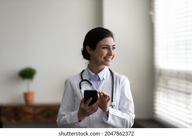 Happy Indian GP doctor chatting online on mobile phone in hospital office, looking at window away, thinking over answer to patient. Young practitioner giving online consultation on smartphone