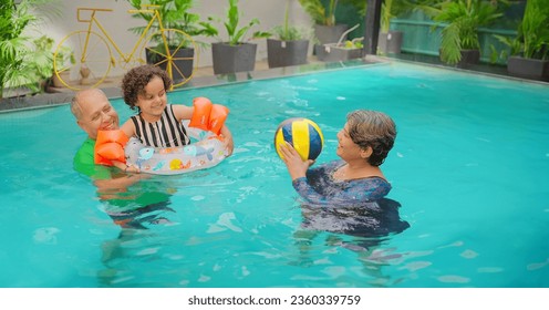 Happy Indian grandparents standing in blue water swimming pool playing ball with cute little girl child outdoor home. Smiling senior couple family enjoying summer vacation holiday in hotel resort  - Powered by Shutterstock