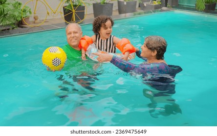 Happy Indian grandparents standing in blue water swimming pool playing ball with cute little girl child outdoor home. Smiling senior couple family enjoying summer vacation holiday in hotel resort  - Powered by Shutterstock