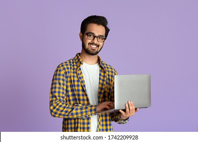 Happy Indian Guy With Laptop Computer Working Or Studying Online On Lilac Background