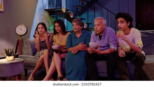 Happy Indian joint family sitting on sofa watching horror thriller TV with scared faces grandparents eating snack young teen girl hold popcorn boul people hanging out together late night film at home  - Powered by Shutterstock