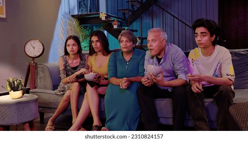 Happy Indian joint family sitting on sofa watching horror thriller TV with scared faces grandparents eating snack young teen girl hold popcorn boul people hanging out together late night film at home  - Powered by Shutterstock