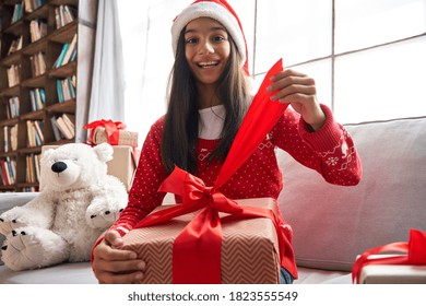 Happy Indian Kid Girl Opening Gift Video Conference Calling Family, Webcam Chat View. Smiling Latin Child Talking To Camera Virtual Meeting Social Distance Online Friend Celebrating Christmas At Home.