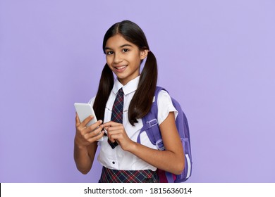 Happy Indian Kid School Girl Holding Phone Standing Isolated On Lilac Background. Smiling Latin Preteen Schoolgirl Wearing School Uniform, Backpack, Using Mobile App On Cellphone Looking At Camera.