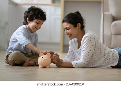 Happy Indian kid and young mom saving money together, putting cash into ceramic piggy bank. Mother playing with child on heating floor at home, teaching little son to invest money, planning future - Shutterstock ID 2074309234