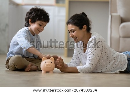 Happy Indian kid and young mom saving money together, putting cash into ceramic piggy bank. Mother playing with child on heating floor at home, teaching little son to invest money, planning future