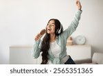 Happy Indian lady with headphones dancing to music and singing song, using hairbrush as mic at home. Young woman moving to her favorite soundtrack, pretending to be rock star