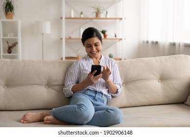 Happy indian lady relax at home alone sit on large couch in comfortable pose share good news at social media via cellphone. Smiling mixed race woman enjoy weekend order goods food online in phone app - Shutterstock ID 1902906148