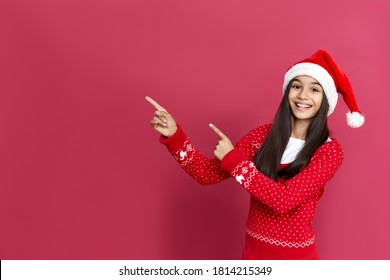 Happy indian latin preteen girl wearing santa hat pointing at copy space standing isolated on red background. Merry Christmas sale, parties for kids, children Happy 2022 New Year celebration concept.
